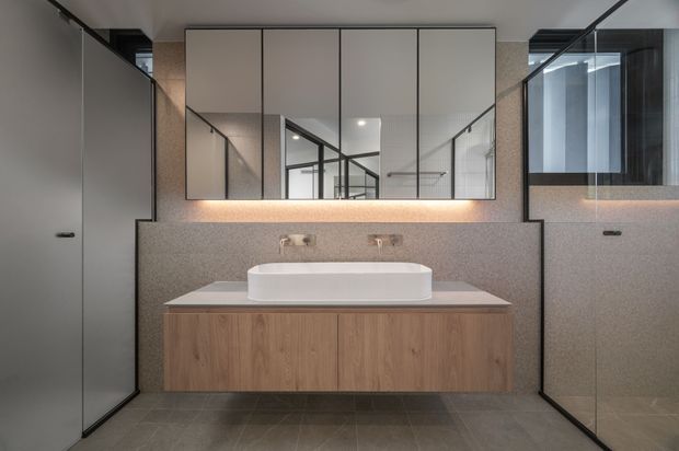 Contemporary bathroom vanity with a floating timber-veneer cabinet, white vessel basin, stone-look benchtop and matching low backsplash, large mirrored wall cabinets, and frameless glass shower enclosures on both sides. The room uses a restrained grey, taupe and warm timber palette with integrated ambient lighting.