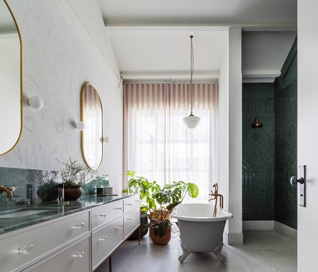 Bright, refined bathroom featuring a freestanding clawfoot bathtub, long double vanity with polished stone surfaces, mirrored wall accents, a dark green tiled shower enclosure, pendant lighting, and sheer curtains over a tall window. The room combines classic fixtures with contemporary and decorative detailing.
