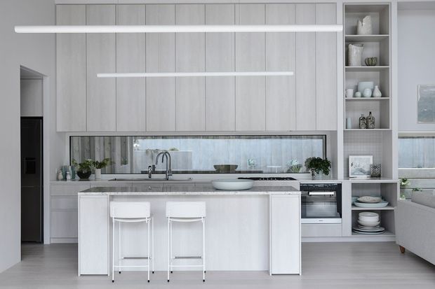 Bright, refined contemporary kitchen with a pale grey-white palette. The space features full-height vertical-panel cabinetry, a long stone-look island with two white stools, integrated shelving displaying crockery, a mirrored or reflective splashback, underbench oven, and minimal linear pendant lighting. A partial living area is visible at the right edge.