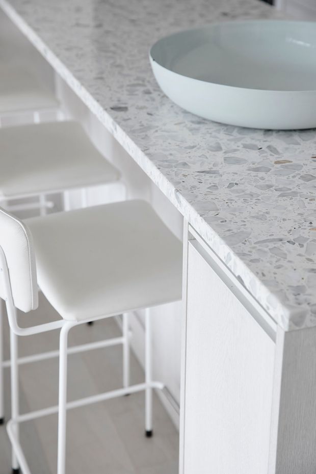 Close-up of a bright, minimalist kitchen or kitchenette featuring a pale terrazzo-style benchtop, white cabinetry, and white bar stools. A white vessel bowl sits on the countertop, with a light neutral palette and clean contemporary detailing.