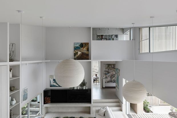 Bright, minimalist contemporary interior viewed across a split-level living space, with white built-in shelving and cabinetry, floating staircases, large spherical pendant lights, high-level glazing with vertical blinds, and artwork. The open-plan arrangement creates a strong sense of layered volume and connection between levels.