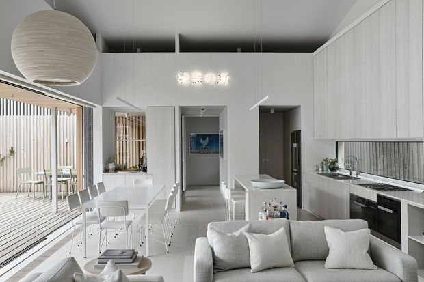 Bright open-plan contemporary interior combining a white kitchen, dining area and living room. The space has extensive white fixed cabinetry, a pale stone-look island and benchtops, light timber flooring, a white dining table with chairs, and a pale upholstered sofa. A glazed or open connection leads to a timber-lined outdoor deck, while a large spherical pendant and compact decorative ceiling light provide feature lighting.