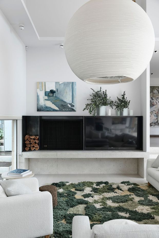 Bright contemporary living room featuring a large sculptural white pendant light, a minimalist concrete fireplace and media wall with dark built-in cabinetry, white upholstered seating, a textured green-and-cream rug, and understated botanical decor. A glazed opening and artwork are visible in the background.