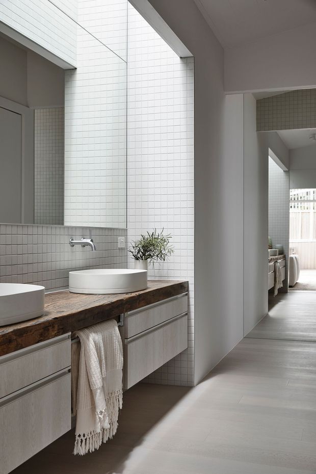 A bright contemporary bathroom with a long floating vanity, two round white vessel basins, wall-mounted chrome tapware, and a thick rustic timber benchtop. Small light-grey grid tiles cover the walls and backsplash, while pale cabinetry and large mirrors create a clean, spacious feel. A hallway or adjacent living area is visible through the opening on the right.