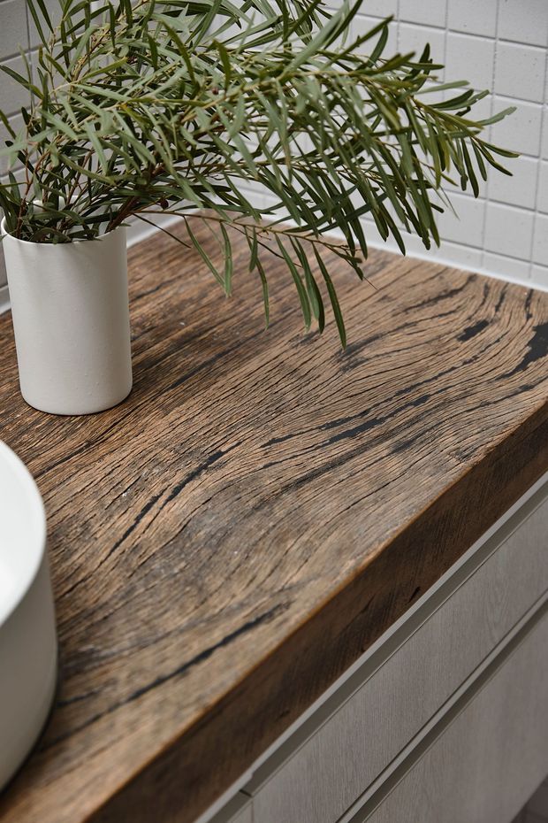 Close-up interior detail of a thick, rustic timber benchtop with prominent dark grain and a live-edge-like textured surface, set above light grey built-in cabinetry. A white cylindrical planter with leafy greenery sits on the counter, with a white tiled wall in the background.