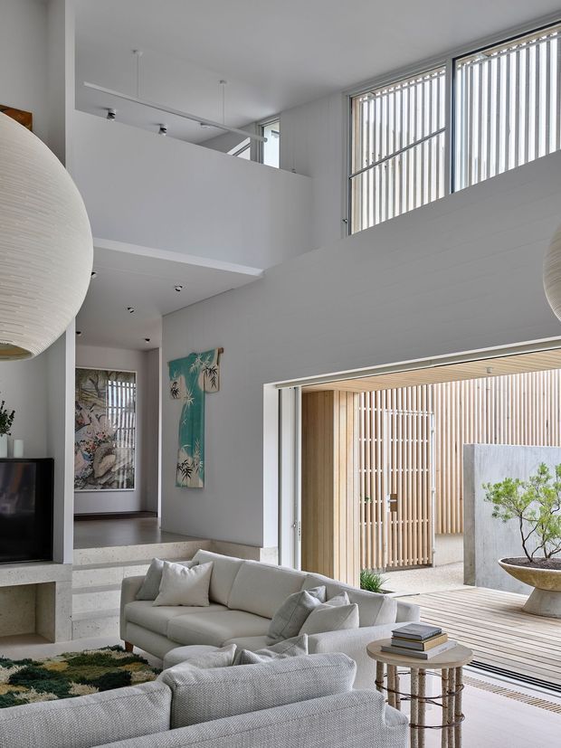 A bright, contemporary double-height living room with a neutral upholstered seating arrangement, large sculptural pendant lights, and a television set into a low built-in wall unit. The room opens directly to a timber-lined courtyard or deck through wide glazed doors, with vertical screening and a planted feature visible outside. High-level glazing with vertical fins, white walls, exposed concrete steps, and a mezzanine-like upper landing create an airy indoor–outdoor connection.