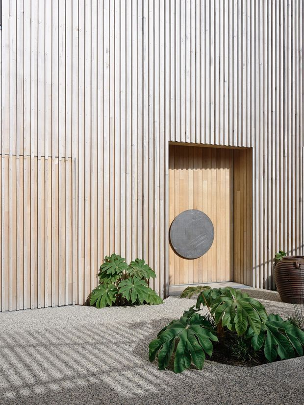 A minimalist courtyard entrance featuring a vertically slatted pale timber-clad facade and recessed timber door. A circular dark metal detail is mounted on the door, with gravel paving, tropical foliage, and a large ribbed planter creating a calm, natural setting. Strong sunlight casts linear shadows across the gravel.