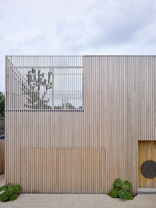 A contemporary building facade clad in closely spaced vertical pale timber battens. The screen-like upper section incorporates an open rectangular window or courtyard opening, while a solid timber-clad wall and recessed entry are visible below. A light concrete or aggregate pavement fronts the facade, with limited planting at the base. The composition is minimalist and strongly vertical beneath an overcast sky.