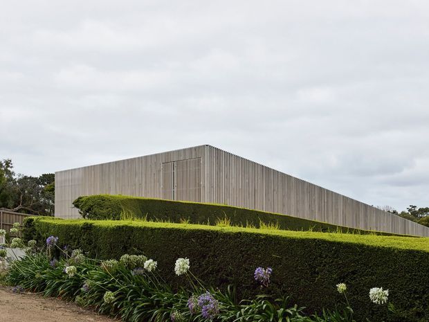 A low, minimalist modern building viewed from the landscaped exterior. The long rectangular form has a flat roof and a facade of closely spaced vertical timber-like battens, creating a screened, textured envelope. Carefully clipped hedge terraces and flowering border planting occupy the foreground beneath an overcast sky.
