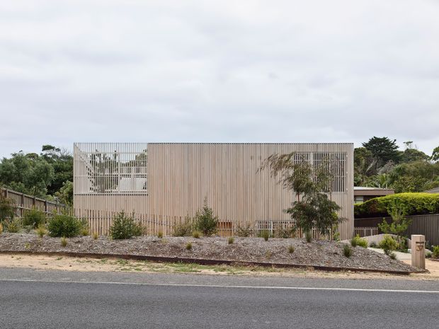 Street-facing view of a compact contemporary residential building with a simple rectangular form, pale vertical timber-like cladding, and extensive white vertical screening over the front glazing and upper openings. The house sits behind a landscaped gravel verge with low planting and boundary fencing, under an overcast sky.