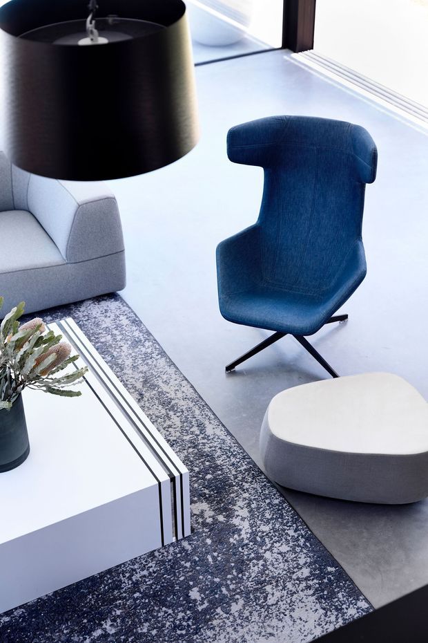 A contemporary living room vignette viewed from above, featuring a sculptural deep-blue upholstered lounge chair, pale grey sofa, rounded light ottoman, large black drum pendant, white geometric coffee table, patterned monochrome rug, and concrete-toned flooring. The scheme is restrained and high-contrast with refined modern furnishings and abundant daylight from large glazing.
