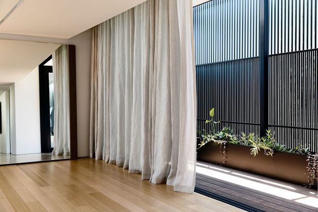 A bright, refined interior with pale timber flooring, full-height sheer linen curtains, and dark-framed glazing opening onto a narrow balcony or terrace. Vertical black screening creates a strong architectural feature outside, while soft natural light and sparse furnishings give the room an airy, minimalist character.