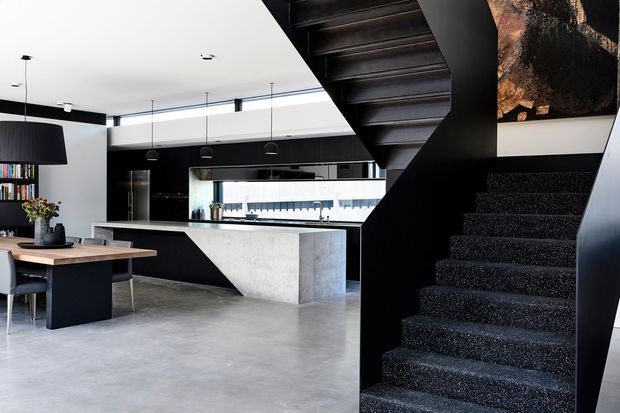 A contemporary open-plan kitchen and dining area in a dark monochrome palette, viewed from beside a dramatic black staircase. The space features a polished concrete floor, black kitchen cabinetry, a large concrete island with a waterfall end, pendant lighting, and a timber dining table. A large artwork and open shelving add visual interest.