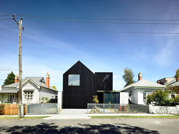 Street-facing view of a contemporary multi-level residence centered between traditional neighboring homes. The house has a steep gabled form clad in dark vertical boards, large black-framed windows, a projecting upper volume, and a low front fence with landscaped planting. Overhead utility lines and a suburban streetscape are visible.