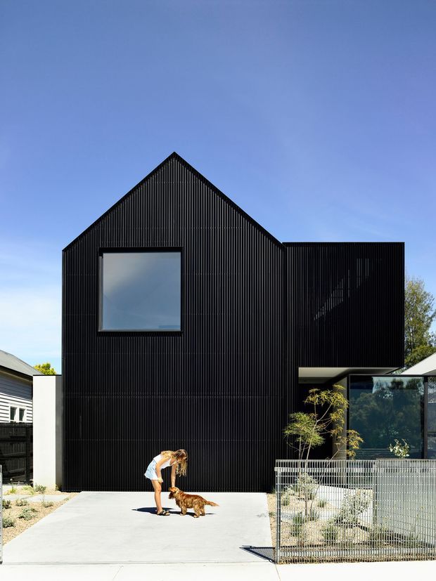 Front exterior view of a contemporary dark-clad residential building with a steep gabled form and a projecting upper volume. The facade is covered in narrow vertical black ribbed cladding, with a large square window and glazed lower-level opening. A concrete driveway, metal front fence, sparse landscaping, neighbouring houses, and a person with a dog are visible.