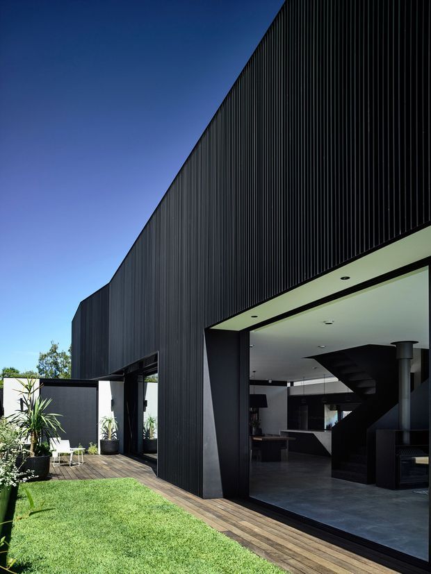 Exterior architectural photograph of a contemporary dark-clad building with a tall, sharply angled roofline and vertically ribbed facade. A large open glazed garage-like opening reveals a dark modern interior with a prominent staircase and kitchen or living area. Timber decking, lawn, and potted plants occupy the foreground.