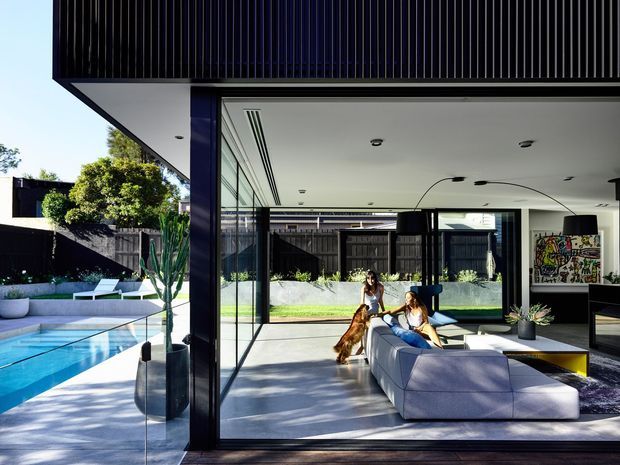 Contemporary open-plan living area viewed through fully glazed sliding doors, with a large sectional sofa, low coffee table, artwork, and indoor plants. The room opens directly onto a swimming pool and landscaped courtyard, with strong indoor-outdoor connection, dark vertical facade cladding, and a projecting flat roof.