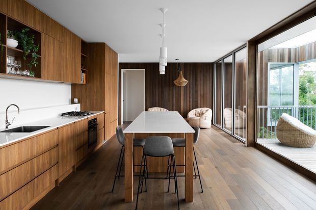 A warm, contemporary open-plan kitchen and dining area with extensive dark timber cabinetry and vertical timber wall panelling. The kitchen includes a white benchtop, integrated appliances and a black sink, while a timber dining table with upholstered stools occupies the centre. Full-height glazed doors open to a balcony or terrace, bringing in abundant natural light. A small seating area with armchairs is visible beyond the dining space.