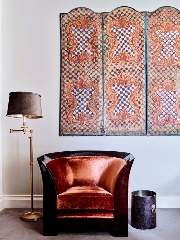 A refined interior vignette featuring a sculptural dark wood or lacquered armchair upholstered in rich rust-red velvet, positioned beside a slim brass floor lamp and a dark metallic wastebasket. A large ornate patterned decorative wall panel in warm terracotta, cream, blue, and brown tones hangs against a pale blue wall with traditional skirting. The composition has a luxurious, dramatic, art-focused character.