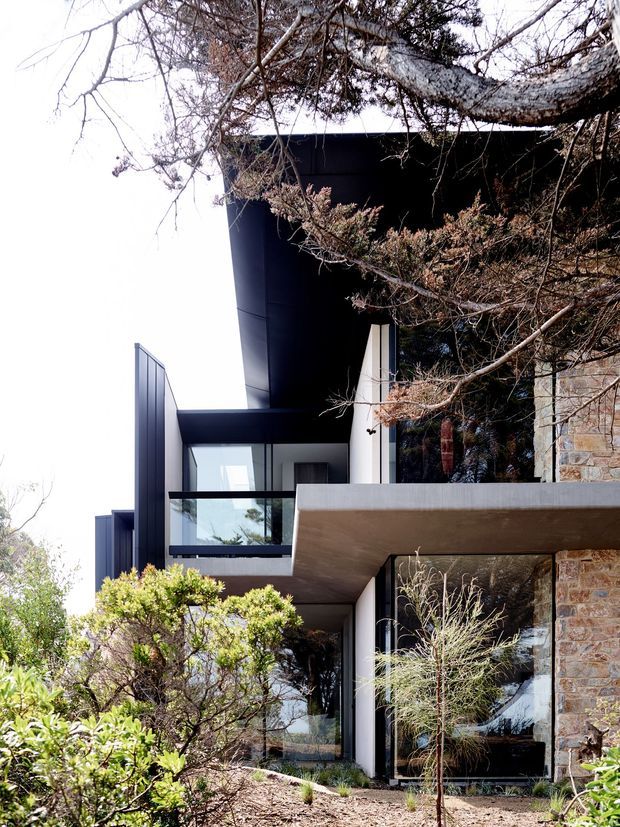 A vertically composed exterior photograph of a contemporary multi-level house integrated into a wooded, rocky site. The building combines dark curved and flat roof forms, sharp cantilevered concrete slabs, extensive glazing with black frames, and textured stone walls. Native-looking planting and mature tree branches partially screen the facade, while balconies and recessed outdoor spaces create layered depth.