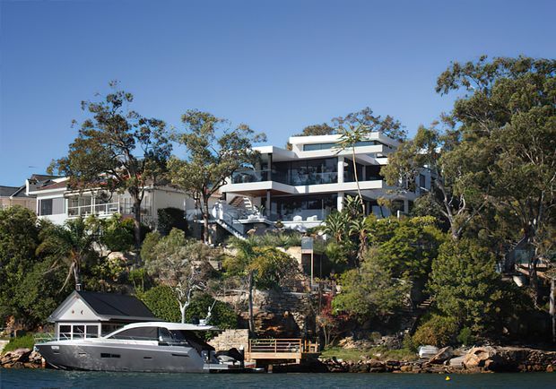 Exterior view of a contemporary multi-level waterfront residence set into a steep, landscaped shoreline. The building features broad glass walls, projecting balconies and terraces, white horizontal slab-like forms, and a flat roof. A private floating dock and large motorboat occupy the foreground, with mature trees and neighbouring homes surrounding the property.
