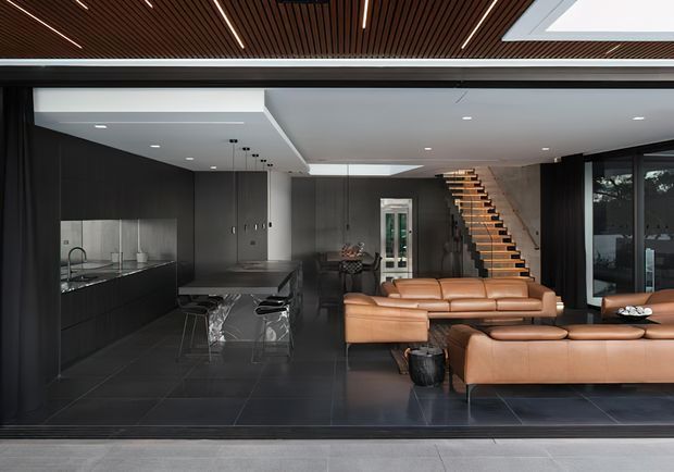 A sophisticated open-plan contemporary interior viewed through a wide glazed opening, combining a dark minimalist kitchen with a central island and stools, a warm tan leather sectional living area, and an open timber stair rising at the rear. Large dark-format floor tiles, charcoal cabinetry and walls, recessed lighting, skylights, and a slatted timber ceiling create a high-contrast, refined atmosphere.