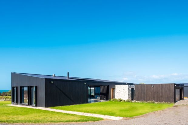 Single-storey contemporary house viewed from the exterior, with a low horizontal form, dark vertical cladding, broad glazing, a pale masonry feature wall, and a flat or very low-pitched roof. The building sits in an open grassy setting beneath a clear sky, with a glimpse of blue water or horizon beyond.