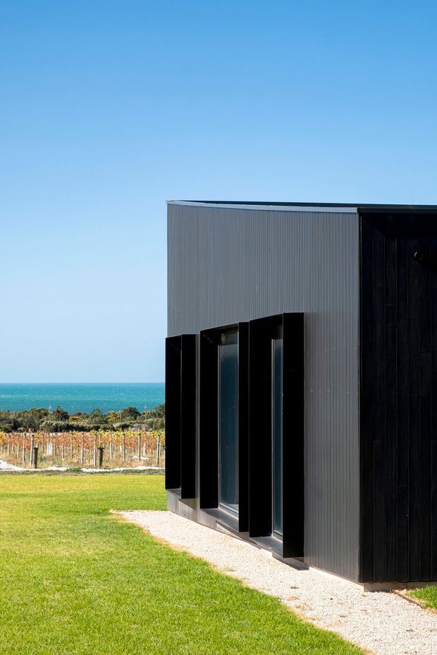 Exterior view of a sleek, box-like coastal building with dark vertical cladding and a light grey corrugated facade. Tall, deeply recessed windows create strong vertical shadow lines. The building sits beside a manicured lawn and gravel path, with a vineyard and turquoise ocean visible beyond under a clear blue sky.