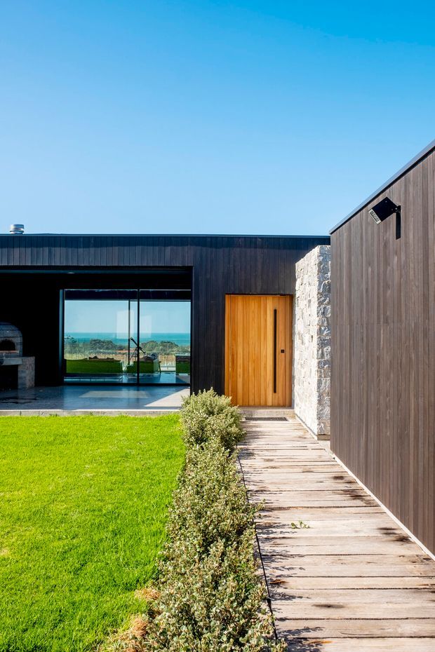 A modern single-storey residence arranged around a landscaped lawn and timber boardwalk. The building has dark vertical cladding, a warm timber entry door, a rough natural-stone wall, broad glazed openings, and a low flat roofline. Through the glazing, a bright coastal or ocean view is visible. The scene is sunny and strongly connected to the outdoor courtyard-like landscape.