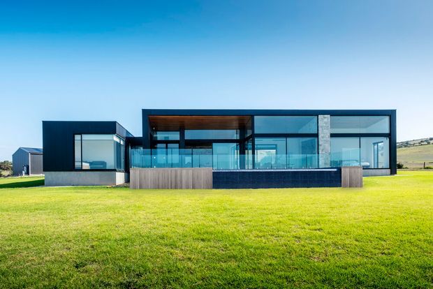 A wide exterior view of a contemporary single-storey house set in an open grassy rural landscape. The building has a strongly rectilinear composition, dark cladding, extensive floor-to-ceiling glazing, a covered central outdoor living area with a timber-lined ceiling, glass balustrades, and a dark masonry or tiled feature wall. A low timber-clad planter or screening element runs along the front terrace.