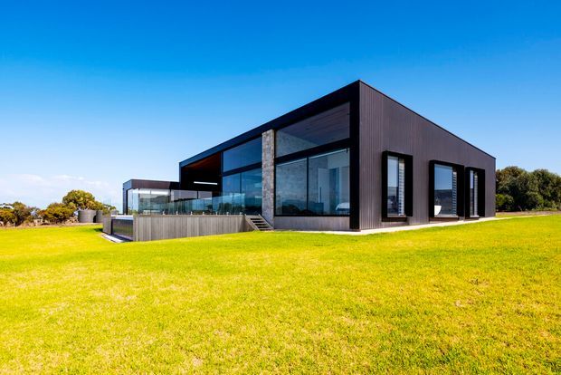 Exterior view of a sleek single-storey modern house set within a broad lawn. The building has a dark vertically ribbed facade, a low asymmetric pitched or folded roof form, extensive floor-to-ceiling glazing, a raised timber-toned terrace, and a prominent stone-clad structural column. The composition is minimalist and strongly connected to the open landscaped site.