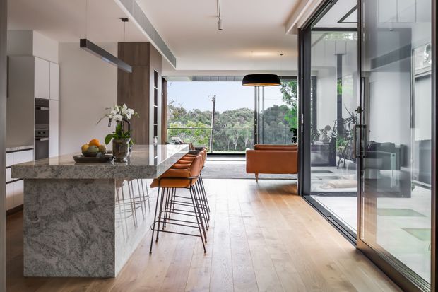 Open-plan contemporary kitchen and living area with a large grey-veined stone island, integrated white cabinetry, timber flooring, and orange-brown bar stools. Full-height black-framed glazing and sliding doors open toward a green outlook, while a leather sofa and pendant lighting define the adjoining living space.
