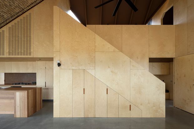 Interior view of a compact open-plan kitchen and living space dominated by a sculptural plywood stair and integrated cabinetry. Pale timber panels form the stair enclosure, storage doors, and wall surfaces, with a darker timber ceiling, exposed ceiling fan, concrete floor, and a partially visible kitchen island. The design is highly streamlined and contemporary, with geometric panel lines and concealed joinery.