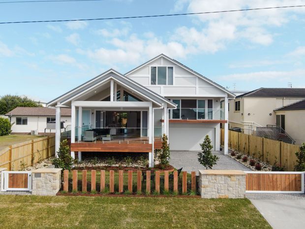 Front exterior view of a multi-level suburban home with a prominent gabled roof, white weatherboard-style facade, covered front porch, timber deck, upper balcony, garage, paved driveway, timber fencing, and low-maintenance landscaping. The home has a bright coastal-influenced appearance with strong gable detailing and open outdoor living areas.