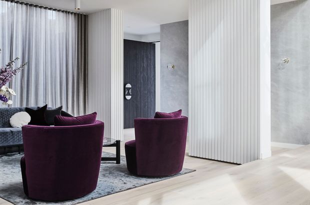 A refined contemporary living room with deep plum velvet lounge chairs and sofa, a black coffee table, layered rugs, sheer floor-to-ceiling curtains, and sculptural fluted white wall panelling. Pale timber flooring, a textured grey feature wall, and a dark vertically panelled doorway create a calm yet luxurious interior.