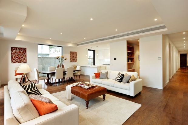 A bright, open-plan apartment interior combining a living room and dining area. Two white sofas surround a large tufted brown leather ottoman on a pale rug, with patterned cushions and orange accents. A dining table sits near large windows, while recessed lighting, integrated air conditioning, timber flooring, white walls, and built-in shelving line the space. A long hallway extends toward the rear.