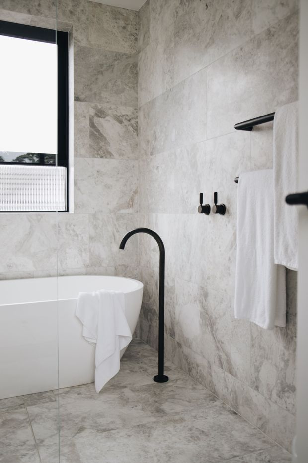 A refined contemporary bathroom featuring a freestanding white bathtub, tall matte-black floor-mounted bath filler, black wall-mounted fixtures and towel rails, and large-format pale grey marble-look wall and floor tiles. A window with a white blind brings in natural light, while white towels add a clean, spa-like feel.