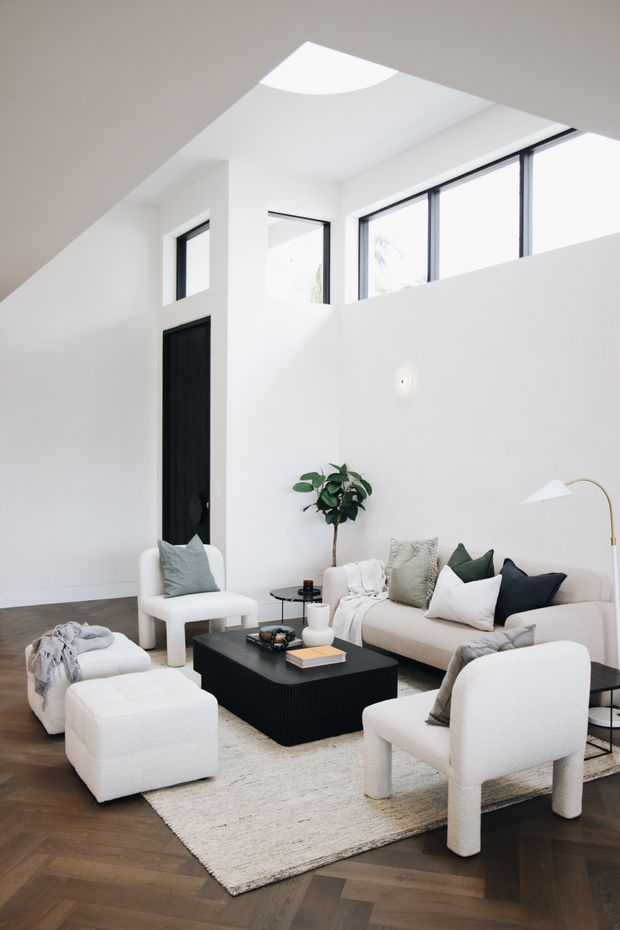 Bright, refined living room with a high ceiling and clerestory-style windows. The predominantly white space features a beige sectional sofa, boucle lounge chairs and ottomans, a black low coffee table, neutral rug, dark timber flooring, black-framed glazing, and a tall indoor plant. A large skylight or roof opening adds abundant natural light.