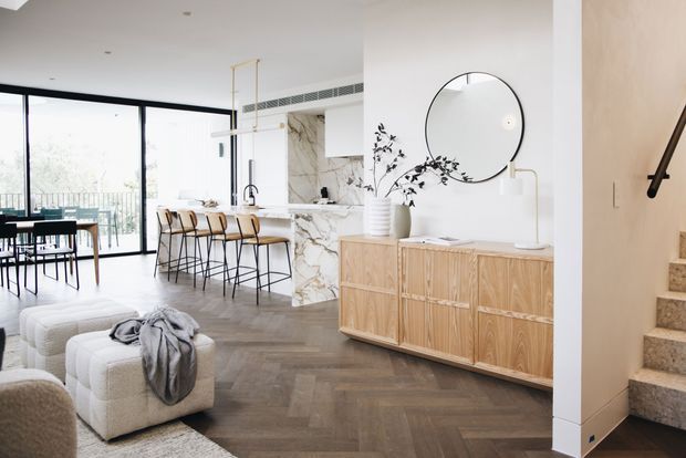 Bright open-plan contemporary interior showing a living area, dining area, and kitchen. The space features floor-to-ceiling black-framed glazing, a marble-clad kitchen island and splashback, light timber cabinetry, herringbone timber flooring, white walls, and a staircase with a black handrail. Neutral upholstered seating and a round wall mirror add softness to the refined, airy scheme.