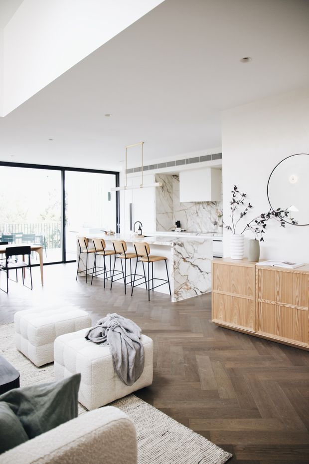 Bright open-plan interior combining a contemporary kitchen, dining area and living room. The space features a large marble-look kitchen island and splashback, white cabinetry, black-framed floor-to-ceiling glazing, pale timber dining furniture, a sculptural linear pendant, light timber sideboard, and dark herringbone timber flooring. Neutral upholstered seating and restrained botanical decor create a refined, airy atmosphere.