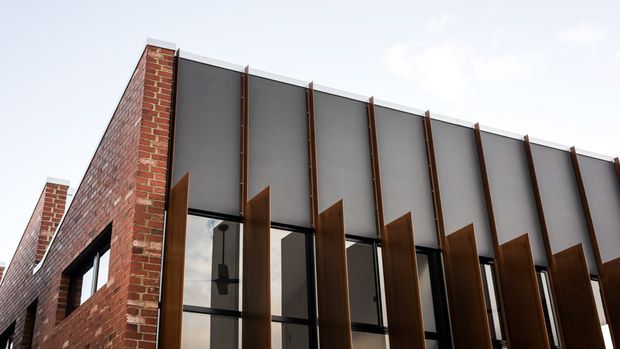 Low-angle exterior view of a contemporary building combining exposed red brick walls with a dark grey metal-clad upper facade. Tall black-framed glazing is screened by a rhythmic series of projecting rust-toned vertical fins, creating a strong modern architectural composition.