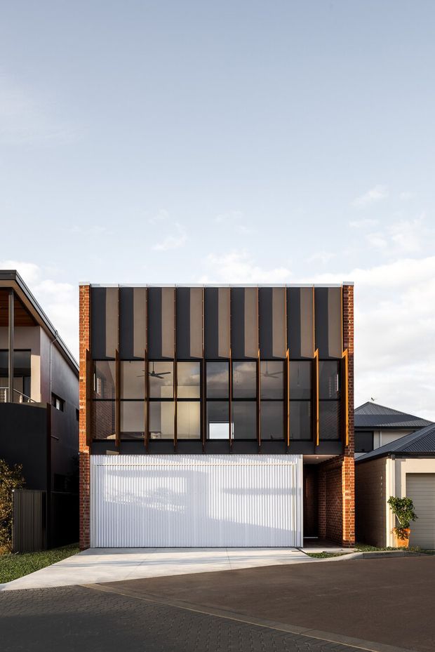 Street-facing exterior view of a contemporary multi-storey townhouse or compact urban residence. The facade combines exposed red brick side walls, a dark metal upper facade with projecting vertical fins, extensive black-framed glazing, and a white ribbed garage door at ground level. Adjacent residential buildings and paved street are visible, with a clear sky above.