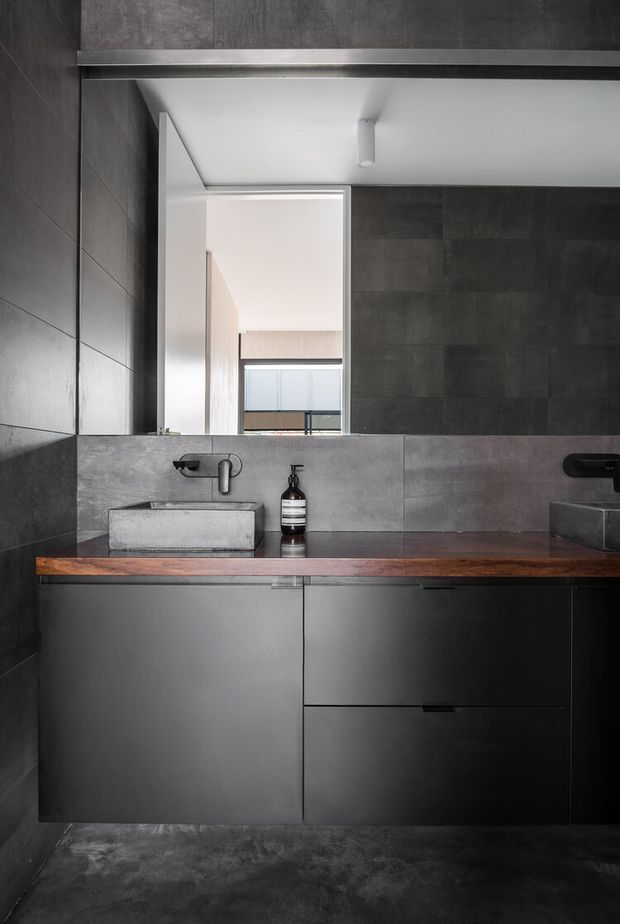 A compact, dark-toned contemporary bathroom vanity with charcoal tiled walls and floor, a rectangular concrete vessel basin, wall-mounted black tapware, dark fixed cabinetry, and a warm timber benchtop. A large mirror reflects an adjacent light-filled interior space, while a horizontal metal trim and concealed lighting add a crisp architectural detail.