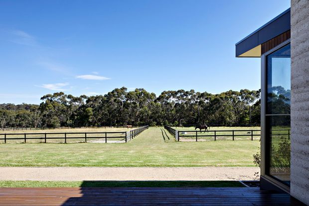 Exterior view from a contemporary building or deck looking across a broad, fenced rural property toward a tree line. The image prominently shows dark timber decking, a sharply projecting flat roof or eave, large dark-framed glazing, and a light textured masonry wall at the right edge. The landscape includes paddocks and fencing, with a strong indoor-outdoor connection and clear blue sky.