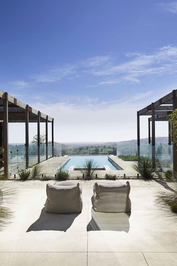 A bright outdoor terrace overlooking a broad rural landscape, centered on a rectangular infinity-edge pool. Two light neutral lounge chairs sit on a pale concrete or stone-paved deck in the foreground. Dark timber or steel pergola frames and clear glass balustrades flank the pool, with sparse ornamental grasses integrated around the edges. The scene has strong sunlight, expansive sky, and a clean indoor-outdoor architectural composition.