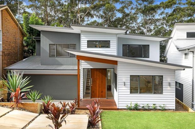 Street-facing view of a contemporary multi-level house with overlapping low-pitched roof planes, white horizontal weatherboard cladding, grey vertical wall panels, dark-framed windows, and a timber-clad recessed entry. A covered carport or garage sits to the left, with a landscaped front garden and concrete path.