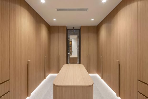 A narrow, highly finished walk-in wardrobe or dressing room lined with full-height warm wood-grain cabinetry. Long vertical handles and low-level integrated lighting define both side walls, while a central wood-clad island with a rounded end provides storage and circulation around it. The room has a smooth white ceiling with recessed downlights and a linear air-conditioning vent, pale carpeted flooring, and a mirrored or glazed opening at the far end. The overall design is symmetrical, calm, and contemporary with strong built-in joinery.