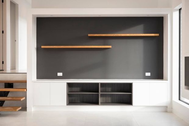 Bright minimalist living room featuring a white built-in entertainment/storage unit, dark charcoal feature wall, open dark timber shelving, and two floating timber shelves. A partial open-riser stairway is visible at left, with large windows providing natural light.