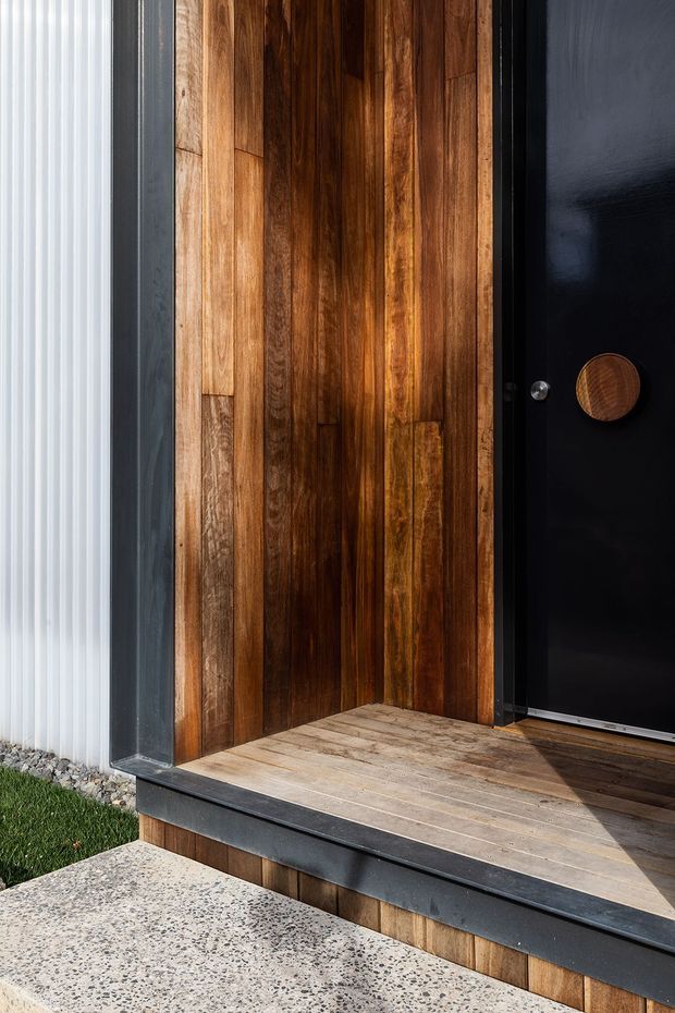 Close-up exterior view of a contemporary entry porch featuring richly stained vertical timber wall cladding, a large matte-black door with a round timber handle, black metal trim, pale timber decking, and a speckled concrete step. White ribbed cladding and a narrow strip of landscaping are visible at the edge.