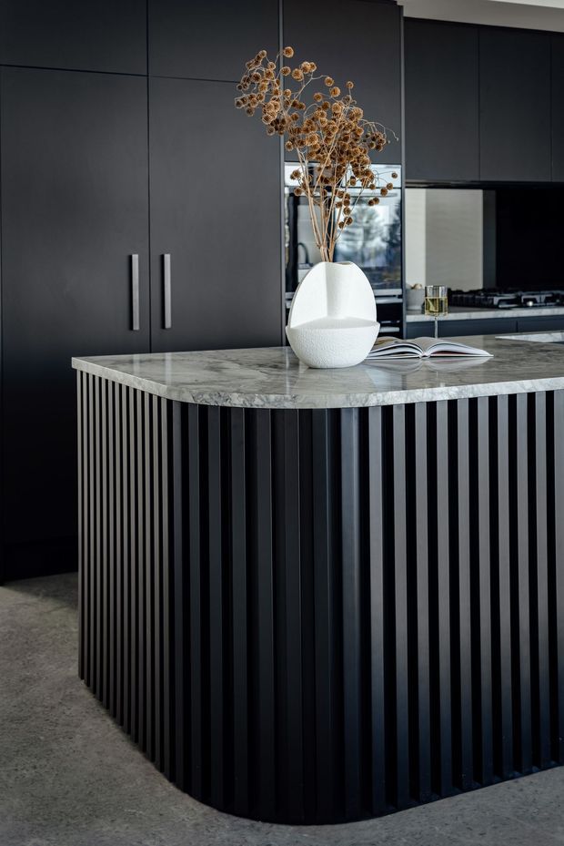 A dramatic, contemporary kitchen centred on a large island with a veined grey stone benchtop and black vertically fluted cabinetry. Full-height matte black kitchen cabinetry, integrated appliances, dark splashback, and a reflective glass or mirrored panel create a sleek monochromatic composition. A sculptural white vase with dried branches and an open book add restrained styling.